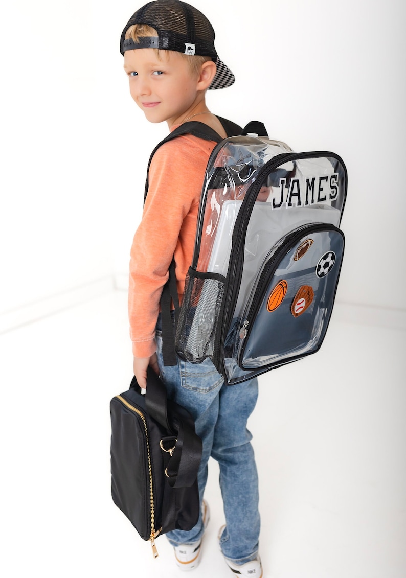May include: A clear plastic backpack with black trim and straps, featuring the name JAMES in white letters and sports-themed patches including a football, soccer ball, basketball, and baseball. A solid black lunch bag is held in the hand.