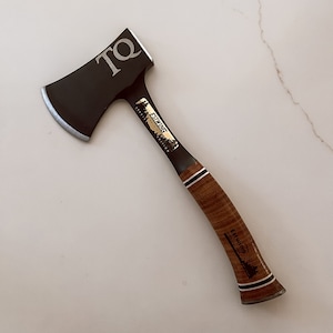 May include: A black hatchet with a silver blade and a brown leather handle. The hatchet has the initials "TQ" on the blade and the brand name "Estwing" on the handle. The hatchet is approximately 14 inches long.