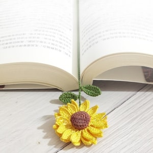 May include: A crocheted sunflower bookmark with green leaves. The sunflower is yellow with a brown center. The bookmark is laying on a white wooden surface.