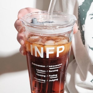 May include: A clear glass tumbler with a lid and straw, filled with a brown beverage and ice cubes. The tumbler has the letters "INFP" in white, along with personality trait pairings. The tumbler is held by a person.