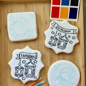 May include: A set of four sugar cookies decorated with stencils for Ramadan and Eid Mubarak. The cookies are on a wooden tray with six colorful paintbrushes. The stencils include a crescent moon, stars, lanterns, and a mosque.