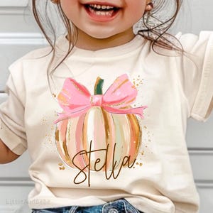 May include: A young child wearing a cream-colored t-shirt with a pumpkin design and the name "Stella" in gold script. The pumpkin has a pink bow and is striped with pink, peach, and gold. The child is wearing sunglasses and blue jeans.