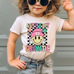 May include: A young child wearing a white t-shirt with the words "A Little Bit Dramatic" and a smiley face graphic. The child is also wearing denim shorts and sunglasses. The shirt features a checkerboard pattern and colorful flowers.
