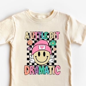 May include: Cream-colored t-shirt with a colorful graphic that says "A LITTLE BIT DRAMATIC." The graphic features a smiley face wearing a pink beanie and peace sign. The shirt is made of 100% cotton.