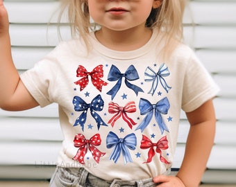 May include: A young girl wearing a white t-shirt with a red, white, and blue bow pattern. The shirt has a crew neck and short sleeves. The girl is also wearing denim shorts and sunglasses.