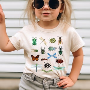 May include: A young child wearing a cream colored t-shirt with a colorful insect print. The shirt has a variety of insects including butterflies, beetles, dragonflies, and ladybugs. The child is wearing denim shorts and sunglasses.
