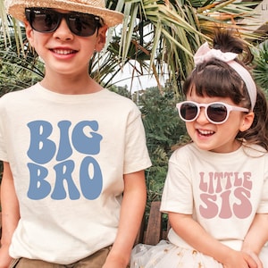 May include: Two children wearing matching t-shirts. The older child is wearing a light blue t-shirt with the text "BIG BRO" in a retro font. The younger child is wearing a pink t-shirt with the text "LITTLE SIS" in a retro font.