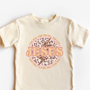 May include: A beige short-sleeved t-shirt with a pink and brown leopard print circle design. The circle has the words "THE WAY THE TRUTH THE LIFE" around the outside and "JESUS" in pink letters inside the circle.