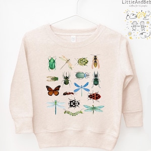 May include: A beige sweatshirt with a colorful insect print. The print features a variety of insects, including butterflies, dragonflies, beetles, and ants. The sweatshirt has a crew neck and long sleeves. The text "LittleAndBebe clothes for little people" is printed on the right side of the sweatshirt.