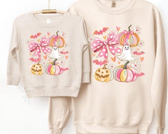 May include: Two beige sweatshirts, one adult and one child size, hanging on a wooden rack. Each features a Halloween-themed design with pumpkins, a ghost, and a pink bow. The text on the image reads: "KIDS AND ADULT SWEATSHIRT COLORS LOOKS EXACTLY SAME. THE HEATHER FABRIC HAS FLECKS IN IT."