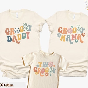 May include: Three t-shirts with a groovy theme. The shirts are beige with colorful text and peace signs. The text on the shirts reads "Groovy Daddy", "Groovy Mama", and "Two Groovy".