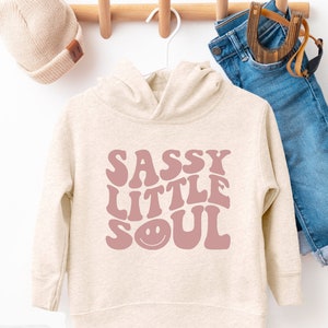 May include: A beige hooded sweatshirt with a pink graphic that says "Sassy Little Soul" with a smiley face.