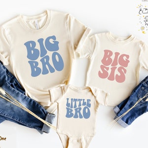 May include: Three cream-colored shirts with blue and pink text. The shirts say "Big Bro", "Big Sis", and "Little Bro" in a retro font. The shirts are laid out on a white surface with blue jeans and dried flowers.