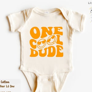 May include: A white baby onesie with a yellow graphic that says "ONE COOL DUDE" in a retro font. The graphic includes two smiley faces with sunglasses.