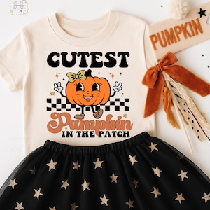 May include: A cream-colored t-shirt with the words "Cutest Pumpkin in the Patch" and a cartoon pumpkin graphic. The outfit includes a black tulle skirt with gold stars and a decorative banner that says "Pumpkin".