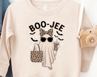 Puede incluir: Sudadera beige con un diseño temático de Halloween. El diseño presenta un fantasma con gafas de sol y un lazo, sosteniendo una bolsa con estampado de leopardo y una bebida. El texto "BOO-JEE" está encima del fantasma. Un peto vaquero está a la izquierda.