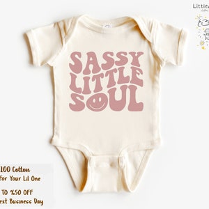 May include: A cream-coloured baby onesie featuring the words "SASSY LITTLE SOUL" in pink, retro-style lettering. The onesie is made from 100% cotton. The brand logo "LittleAndBebe" is in the upper right corner.