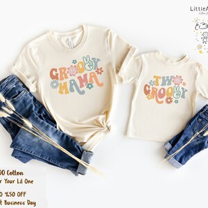 May include: Two matching t-shirts with a groovy theme. The larger shirt says "Groovy Mama" in colorful letters with peace signs and flowers. The smaller shirt says "Two Groovy" in colorful letters with peace signs and flowers.