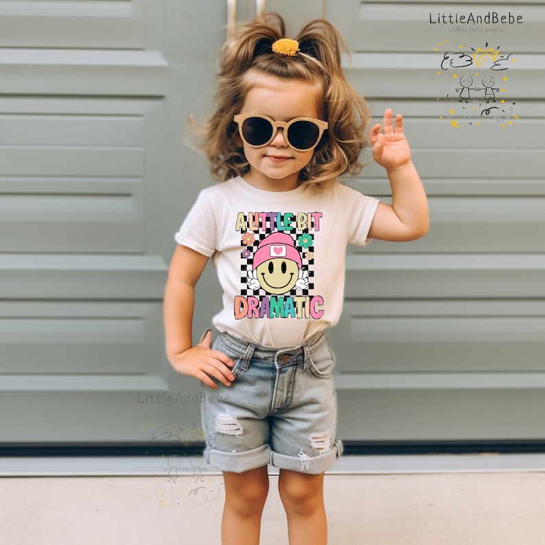 May include: A young child wearing a white t-shirt with the words "A Little Bit Dramatic" and a smiley face graphic. The child is also wearing denim shorts and sunglasses. The shirt features a checkerboard pattern and colorful flowers.