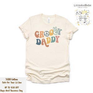 May include: A beige t-shirt with the text "Groovy Daddy" in rainbow colors. The text includes a peace sign and a peace hand gesture.