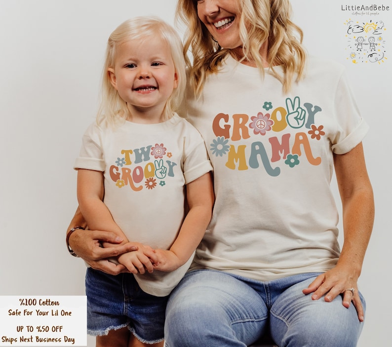 May include: A mother and child wearing matching white t-shirts with a groovy theme. The mother's shirt says "Groovy Mama" and the child's shirt says "Two Groovy". Both shirts feature a peace sign and flowers in a retro style.