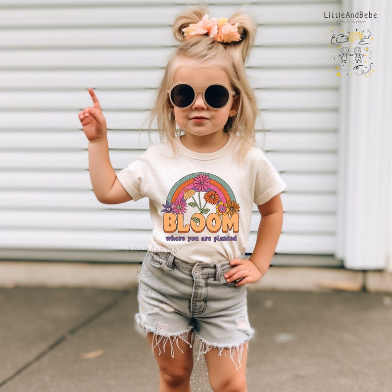 May include: A young girl wearing a light beige t-shirt with a rainbow and flower graphic and the text "Bloom where you are planted". She is wearing dark sunglasses and denim shorts.