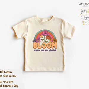 May include: A cream colored t-shirt with a rainbow and flowers graphic. The text "BLOOM where you are planted" is printed on the shirt.