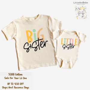 May include: Two matching cream-colored children's outfits. One is a t-shirt with "BIG sister" in colorful letters and black script. The other is a onesie with "LITTLE sister". The text "%100 Cotton Safe For Your Lil One" is also visible.