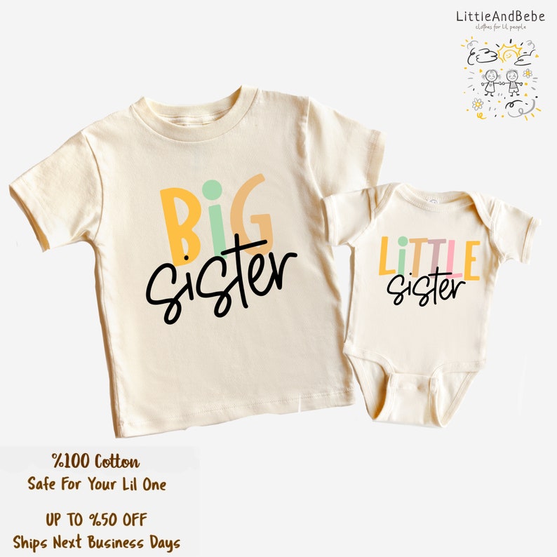 May include: Two matching cream-colored children's outfits. One is a t-shirt with "BIG sister" in colorful letters and black script. The other is a onesie with "LITTLE sister". The text "%100 Cotton Safe For Your Lil One" is also visible.