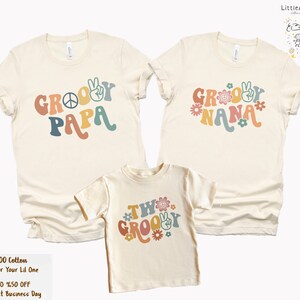 May include: Three cream-colored t-shirts with a retro groovy font design. The shirts say "Groovy Papa", "Groovy Nana", and "Two Groovy". Each shirt has a peace sign and flowers.