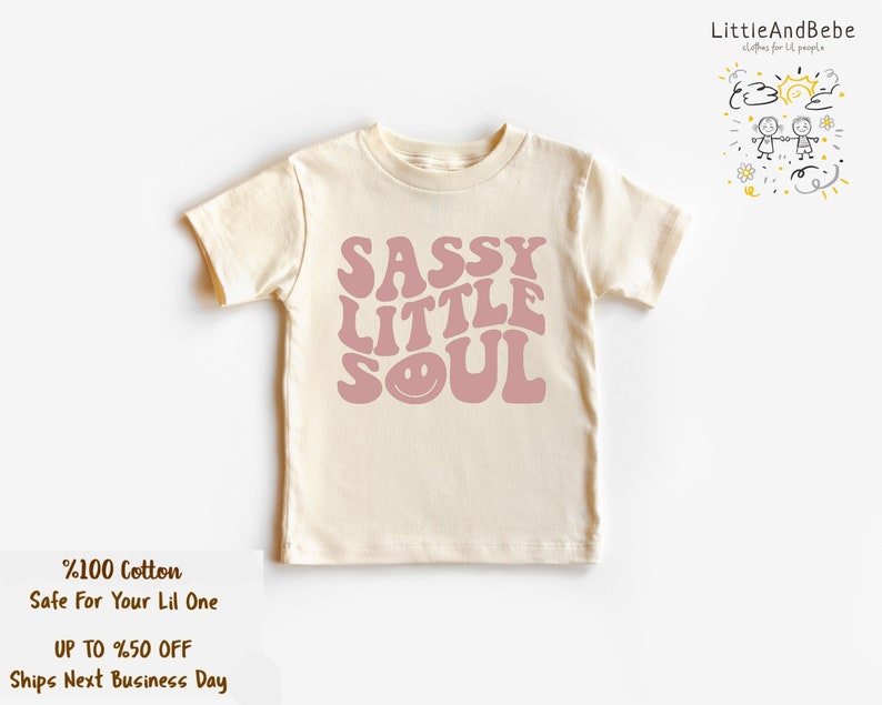 May include: A cream-coloured t-shirt featuring the phrase "SASSY LITTLE SOUL" in a retro font, with a smiling face graphic. The shirt is made of 100% cotton. The brand logo "LittleAndBebe" is in the upper right corner.
