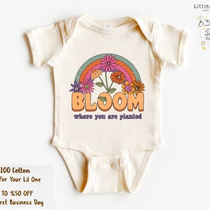 May include: A cream-colored baby onesie with a rainbow and flowers graphic. The text "BLOOM where you are planted" is printed on the onesie.