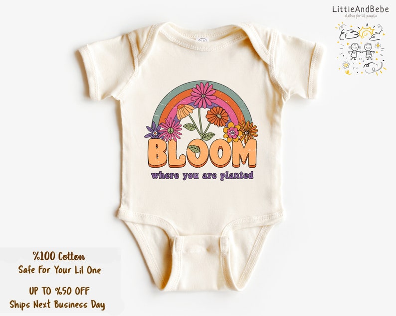 May include: A cream-colored baby onesie with a rainbow and flowers graphic. The text "BLOOM where you are planted" is printed on the onesie.