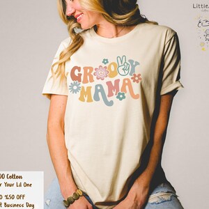 May include: A light beige t-shirt with the text "Groovy Mama" in colourful retro lettering with peace signs and flowers.