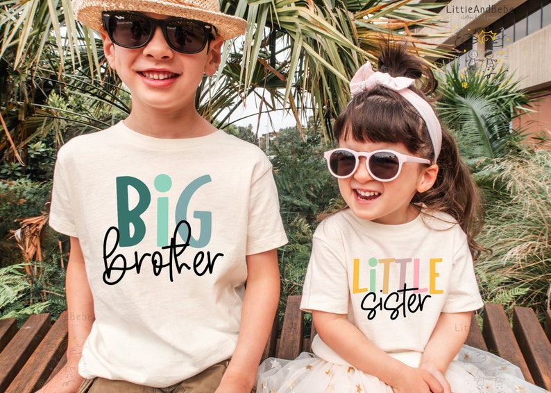 May include: Two children wearing matching cream-colored t-shirts with text. One shirt says "BIG brother" in teal and black, the other says "LITTLE sister" in pastel colors and black. Both children are wearing sunglasses and smiling.