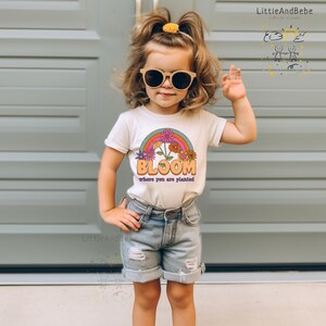 May include: A white t-shirt with a rainbow and flowers graphic. The text "Bloom where you are planted" is printed on the shirt.