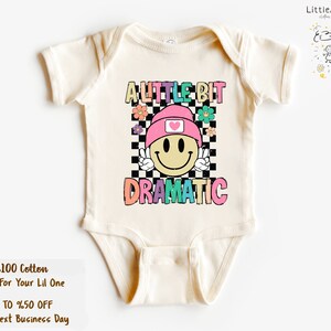 May include: Cream-colored baby bodysuit with the text "A Little Bit Dramatic" in colorful letters. The design features a smiley face wearing a pink hat with a heart, set against a checkered background, and surrounded by flowers. The bodysuit is made of 100% cotton.