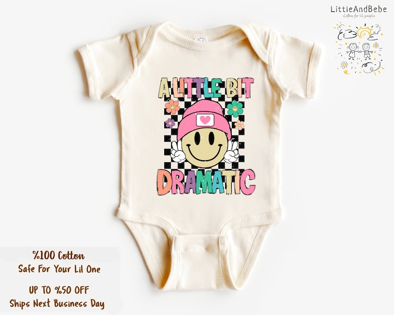 May include: Cream-colored baby bodysuit with the text "A Little Bit Dramatic" in colorful letters. The design features a smiley face wearing a pink hat with a heart, set against a checkered background, and surrounded by flowers. The bodysuit is made of 100% cotton.