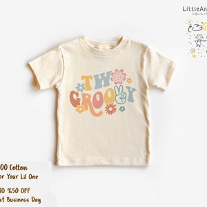 May include: A cream colored short-sleeved t-shirt with the text "Two Groovy" in colorful retro font with peace signs and flowers.