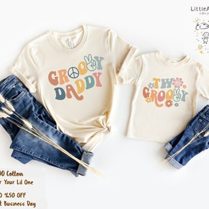 May include: Two matching t-shirts with a retro groovy design. The larger shirt says "Groovy Daddy" in rainbow colors with a peace sign. The smaller shirt says "Two Groovy" in rainbow colors with a peace sign and flowers.