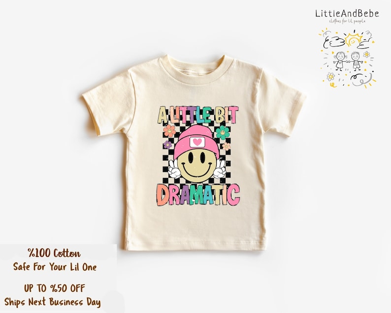 May include: Cream-colored t-shirt with a colorful graphic that says "A LITTLE BIT DRAMATIC." The graphic features a smiley face wearing a pink beanie and peace sign. The shirt is made of 100% cotton.