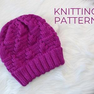 Può includere: Primo piano di un cappello a maglia di colore magenta vibrante. Il cappello presenta un motivo a treccia lavorato a maglia con una texture. Il cappello è appoggiato su una superficie bianca e soffice. Il testo "KNITTING PATTERN" è visibile nell'angolo in alto a destra dell'immagine.