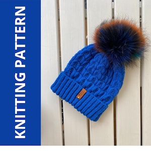 May include: A blue knitted hat with a cable knit pattern and a faux fur pom pom. The hat has a brown leather label with the word "Cozy" on it.