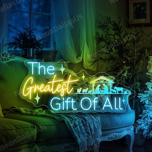 May include: A neon sign with the text "The Greatest Gift Of All" in yellow and white, with a nativity scene silhouette. The sign is on a green couch in front of a Christmas tree with blue and green lights.