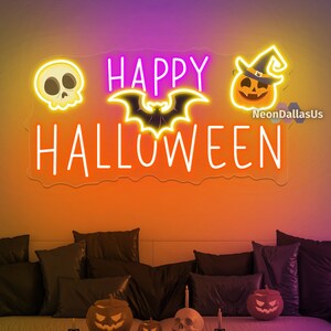 Op de afbeelding: Een neonbord met de woorden "HAPPY HALLOWEEN" in het wit, met schedel-, vleermuis- en pompoen-iconen. Het bord hangt aan een oranje muur boven een bank met zwarte kussens en Halloween-decoraties.
