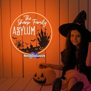 May include: A neon sign with an orange glow, depicting a spooky Halloween scene with a haunted house, bats, and a graveyard. The sign reads "The Sharp Family ASYLUM" with the text "NeonDallasUs" below. A young girl dressed as a witch sits on a wooden floor with a carved pumpkin in front of her.