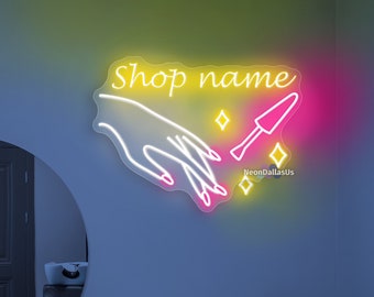 May include: Neon sign featuring a white outline of two hands holding a nail polish bottle and a brush. The sign reads "Shop name" in yellow and pink neon. The sign is mounted on a light blue wall.