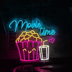 Op de afbeelding: Neon-bord met de woorden "Movie Time" in blauw en roze. Het bord toont ook een cartoon-afbeelding van een emmer popcorn en een glas frisdrank met een rietje en een hart.