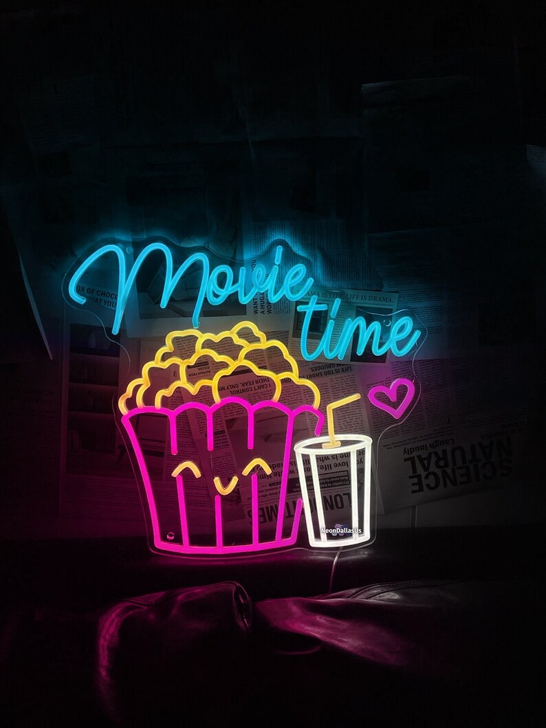 Op de afbeelding: Neon-bord met de woorden "Movie Time" in blauw en roze. Het bord toont ook een cartoon-afbeelding van een emmer popcorn en een glas frisdrank met een rietje en een hart.