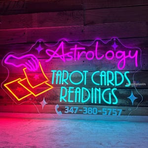 May include: Neon sign with a pink "Astrology" and turquoise "Tarot Cards Readings" text. A hand holding two cards with stars is depicted. The phone number 347-380-5757 is also included.